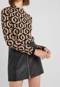 Beżowy sweter z czarnym okrągłym wzorem logo i długimi rękawami, połączony z czarną skórzaną spódnicą z bocznym zamkiem.