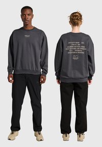 Voor- en achterkant van twee personen die donkergrijze sweatshirts met tekstlogo's en zwarte broeken dragen, staand op een effen lichtgrijze achtergrond.