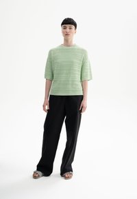 Lichtgroen gebreid T-shirt met korte mouwen en horizontale gestreepte textuur, gecombineerd met zwarte wijde broek en grijze sandalen op een effen achtergrond.