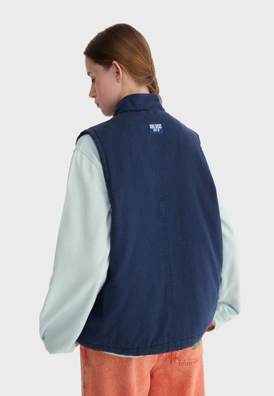 Personne aux cheveux bruns attachés en queue de cheval, portant un gilet bleu marine sur une chemise à manches longues bleu clair et un pantalon orange, dos à la caméra.