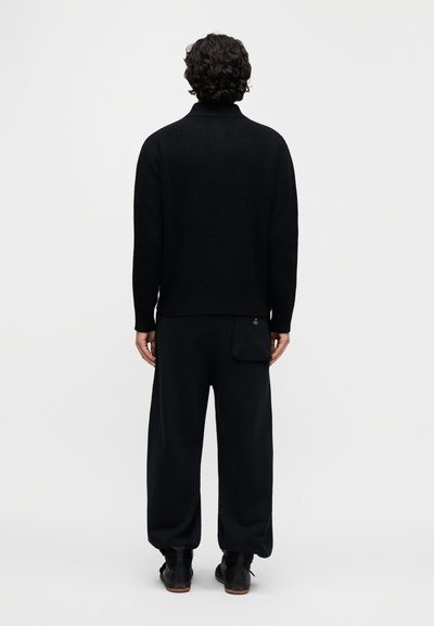 Μαύρο πλεκτό πουλόβερ με ψηλό γιακά, συνδυασμένο με φαρδιά μαύρα παντελόνια με πλαϊνή τσέπη. Ομαλή υφή, ελάχιστες λεπτομέρειες.