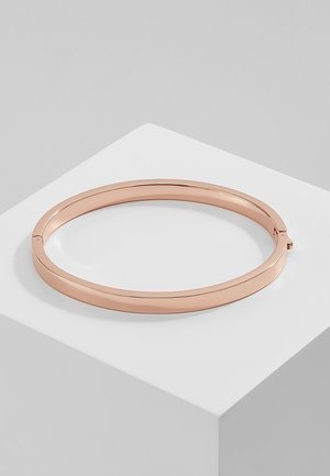 Bracelet jonc en métal couleur or rose avec une finition lisse et polie reposant sur un socle cubique blanc.