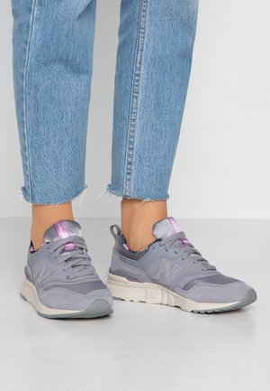 Noha v šedých a růžových teniskách New Balance, spárovaných s modrými džínami s roztřepeným lemem, na světle šedé podlaze.