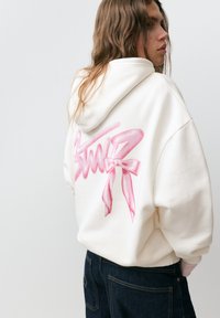 Persoană purtând un hanorac alb cu text roz în stil graffiti și o fundă pe spate, asortat cu blugi albastru închis, pe un fundal simplu.