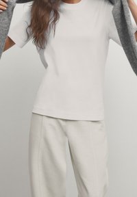 Witte katoenen t-shirt met korte mouwen, ronde hals en relaxte pasvorm, gecombineerd met lichtbeige broek, een grijze cardigan vasthoudend.