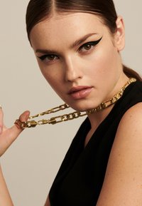 Kultaketju kaulaketju, jota pidetään kädessä, jossa on suuret lenkit. Mallilla on suorat hiukset ja voimakas musta eyeliner, ja hänellä on yllään musta vaate.