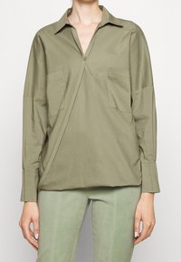 Camicia lunga olive green con colletto a bottoni, due tasche frontali e vestibilità comoda, realizzata in tessuto morbido con una leggera lucentezza.