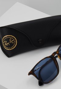 Óculos de sol Ray-Ban com lentes azul-escuras, armações tartaruga e uma caixa preta texturizada com um logótipo dourado.