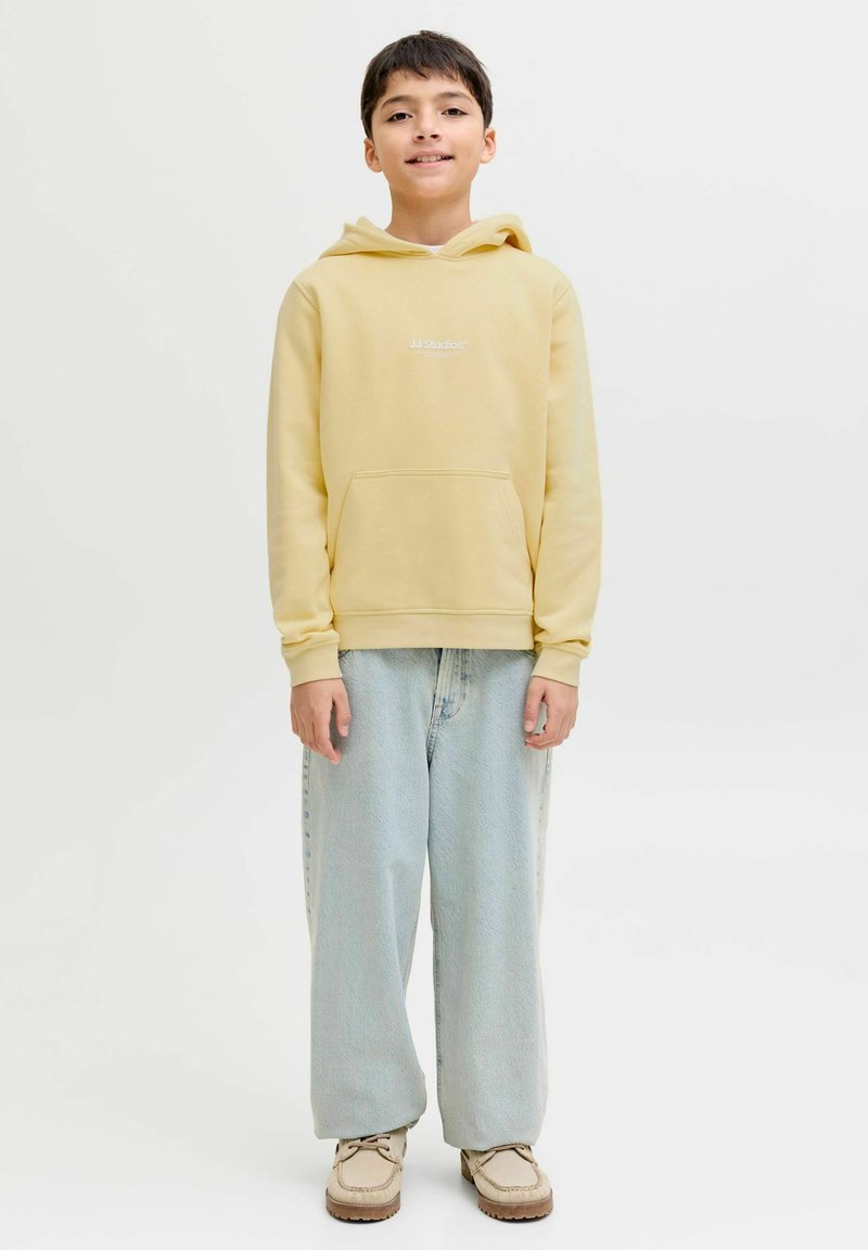 Sweat à capuche en coton jaune avec poche avant, arborant le logo "JJ Studios". Associé à un jean large bleu clair et des baskets beiges.