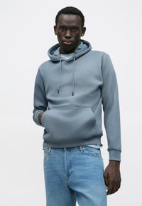 Grå hættetrøje i blødt materiale med en frontlomme, justerbar snøre og ribkant ved ærmerne, kombineret med lyseblå denimjeans.