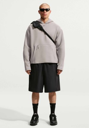 Uomo che indossa una felpa grigia con cappuccio, pantaloncini neri, calzini neri, scarpe da ginnastica nere, occhiali da sole e una borsa a tracolla nera su sfondo semplice.