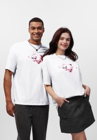 Hvide bomulds T-shirts med korte ærmer, prydet med et pink hjerte-design og ordet "TRUST" trykt i sort, parret med mørke bukser og en nederdel.