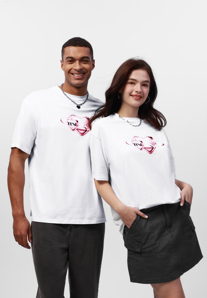 Hvide bomulds T-shirts med korte ærmer, prydet med et pink hjerte-design og ordet "TRUST" trykt i sort, parret med mørke bukser og en nederdel.