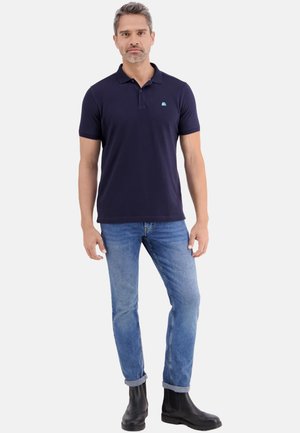 Uomo in piedi che indossa una polo blu navy, jeans azzurri risvoltati e stivaletti neri su uno sfondo bianco neutro.