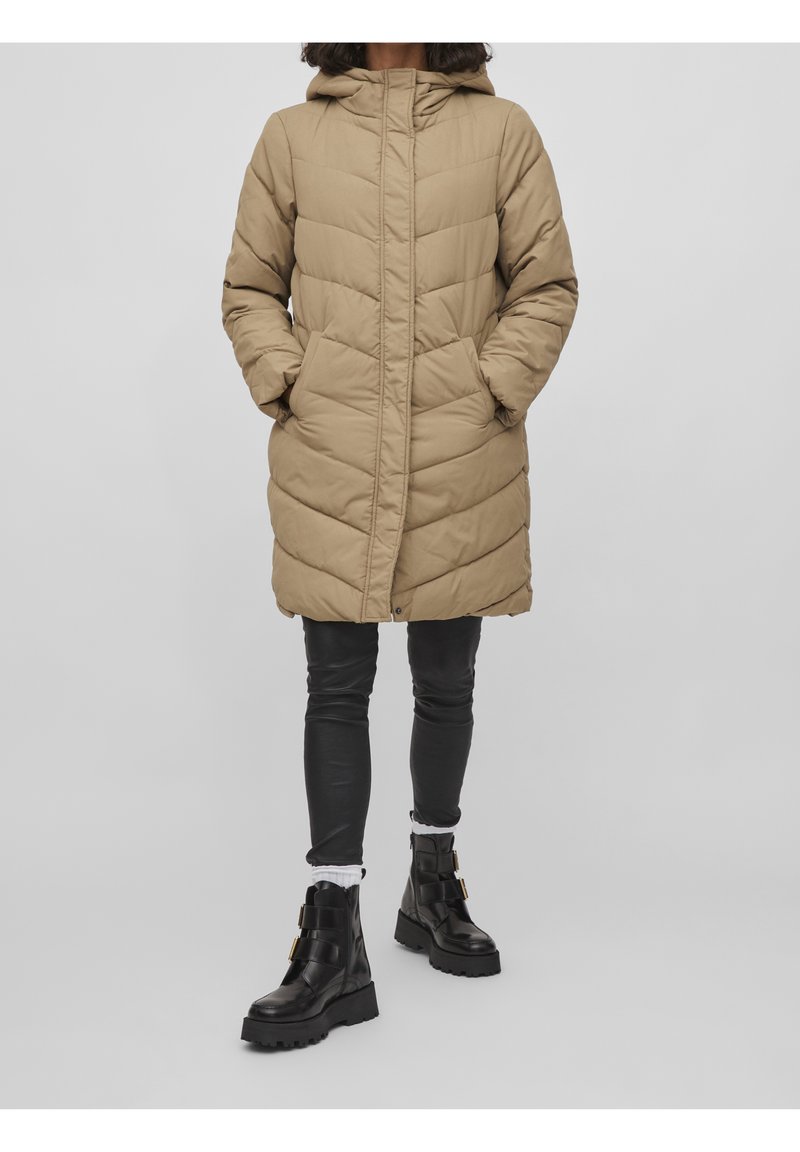 Femme portant un manteau matelassé beige mi-mollet avec capuche, pantalon en cuir noir, chaussettes blanches et bottes noires épaisses, debout devant un fond uni.