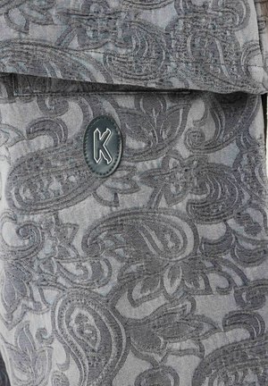 Tela gris con complejos patrones de paisley y un parche ovalado negro con una letra "K" blanca en el centro.
