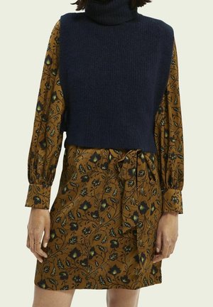 Femme portant une robe marron à fleurs à manches longues avec une taille nouée et un gilet sans manches bleu marine à col roulé en maille.