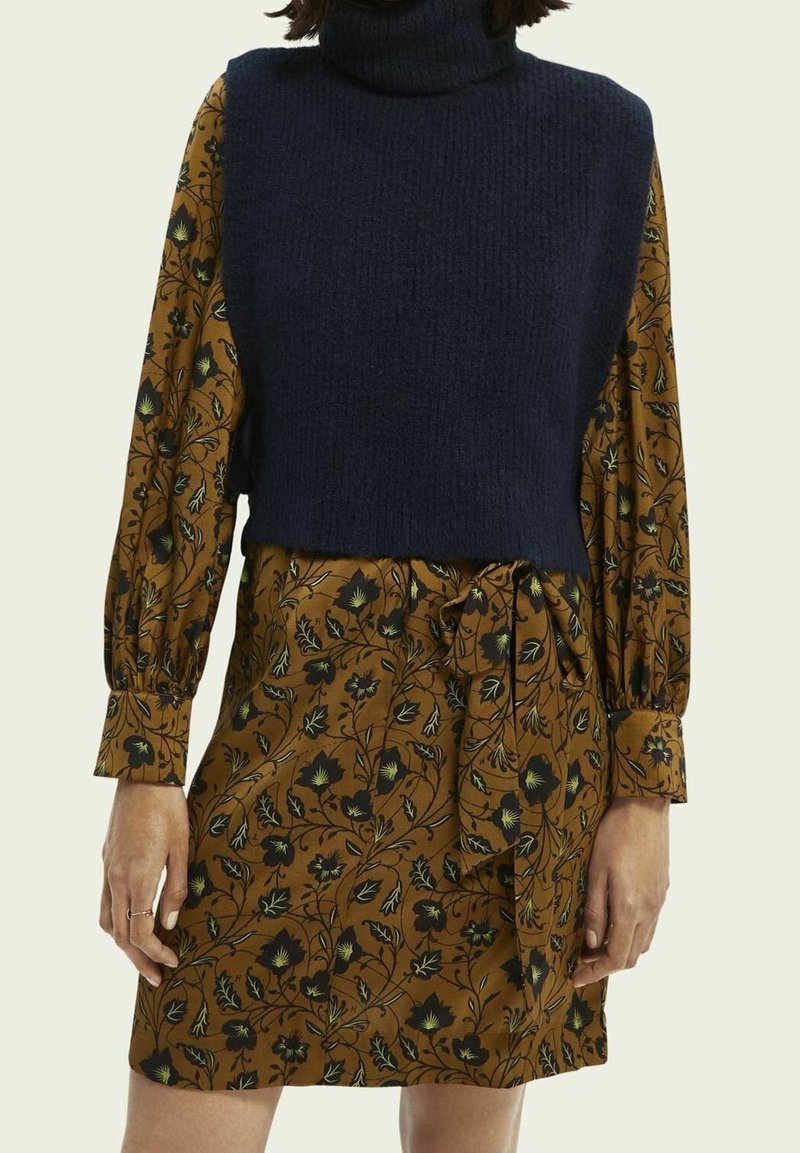 Femme portant une robe marron à fleurs à manches longues avec une taille nouée et un gilet sans manches bleu marine à col roulé en maille.