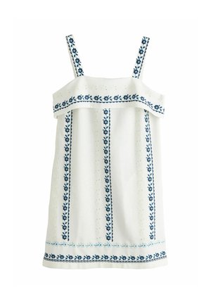 Femme aux longs cheveux foncés dans une robe blanche sans manches avec une broderie florale bleue, appuyée sur un balcon blanc dominant la mer.