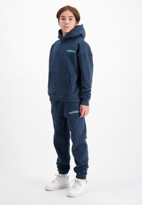 Marineblauwe sweatshirt met verstelbare capuchon, voorkeurszak en groen logo. Gecombineerd met bijpassende broeken, taps toelopende boorden en witte sneakers.