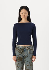 Tamsiai mėlyna ilga rankovė bluzonė su plačiu kaklu, prigludusi liemenį ir be siūlių manžetėmis, derinama su raštuotomis džinsinėmis kelnėmis, puoštomis rudos spalvos gėlių motyvais.