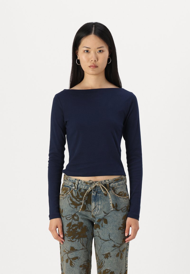 Tamsiai mėlyna ilga rankovė bluzonė su plačiu kaklu, prigludusi liemenį ir be siūlių manžetėmis, derinama su raštuotomis džinsinėmis kelnėmis, puoštomis rudos spalvos gėlių motyvais.