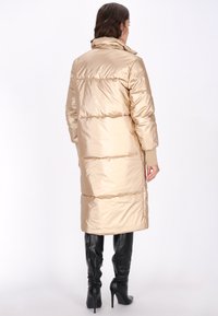 Lang, beige vinterjakke med en blank finish, med høj krave, ribbede manchetter og vandrette quiltede sektioner, der strækker sig til midt på læggen.