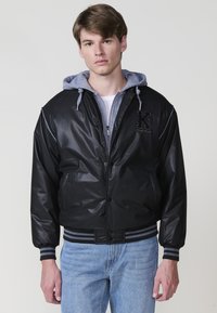 Giacca bomber nera con finitura lucida, sottogiacca grigia con cappuccio, polsini e orlo a costine a righe, e dettaglio ricamato con lettera sul petto.