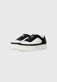 Zwart-witte lage sneakers met vetersluiting en dikke witte zolen, iets naar rechts gekanteld op een effen lichte achtergrond.