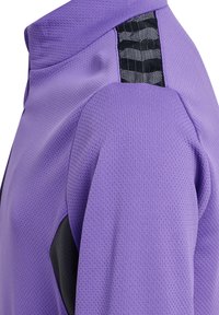 Lila sportjacka med en strukturerad, andningsbar yta, svart accentpanel och unik mönsterdetalj längs axelsömmen.