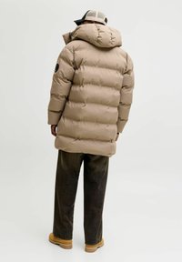 Mies, jolla on beige toppatakki hupulla, tummat housut, ruskeat saappaat ja beige baseball-lippis, seisoo selin valkoisella tasaisella taustalla.