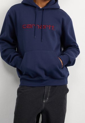 Personne portant un sweat à capuche bleu marine avec le logo rouge "carhartt", les mains dans la poche avant, associée à un pantalon noir sur fond blanc uni.
