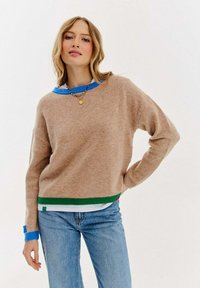 Sweter dzianinowy w miękkim beżu z niebieskim kołnierzykiem i zielonym wykończeniem na dole. Posiada długie rękawy i luźny krój. Założony na biały koszulę.