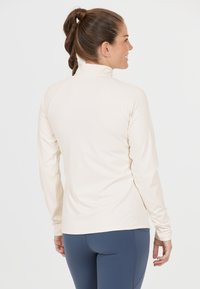 Helles beiges Langarmshirt mit hohem Kragen, glatter Textur und Raglanärmeln. Getragen mit eng anliegenden navyblauen Hosen, von hinten betrachtet.