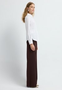 Donna con capelli ricci che indossa una camicia bianca con bottoni, pantaloni a righe marroni a gamba larga, scarpe beige con punta e orecchini a cerchio argentati, in piedi di lato.