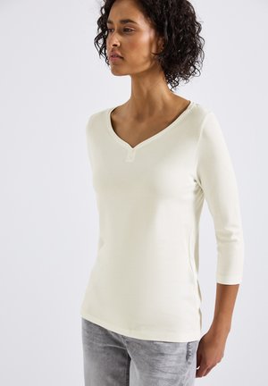 Haut à manches longues crème clair en tissu doux ; il présente un col en V et deux boutons au niveau du col. Associé à un jean gris, révélant une silhouette ajustée.