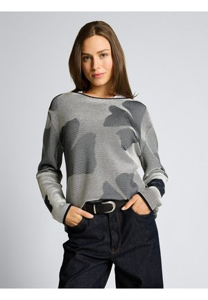 Kobieta o długich brązowych włosach, ubrana w czarno-biały sweter w paski z abstrakcyjnymi wzorami, czarny pasek i ciemne dżinsy, ręce w kieszeniach.