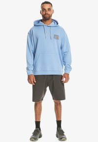 Light blue pullover hoodie with a front pocket and drawstring hood, paired with gray shorts and dark sneakers. Subtle logo on chest.