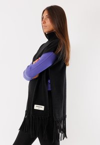 Poncho nero in maglia con colletto alto, caratterizzato da un orlo frangiato e un'etichetta bianca. Sotto, è visibile una maglietta a maniche lunghe viola.