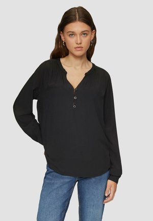 Zwarte longsleeve t-shirt met een V-hals, drie knopen bij de sluiting en geplooide details op de schouders. Zacht materiaal, relaxed fit. Gecombineerd met blauwe jeans.