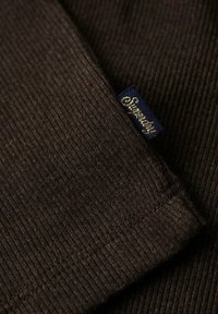 Ciemnobrązowy rypsowy materiał o miękkiej strukturze, z niebieską metką z napisem "Superdry" złotą czcionką.