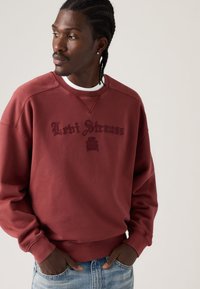 Μπορντό φούτερ με το λογότυπο «Levi Strauss» σε σκούρο κόκκινο. Στρογγυλή λαιμόκοψη, ριμπ μανσέτες και ίσια τελείωμα. Φοριέται με ανοιχτόχρωμο μπλε τζιν.