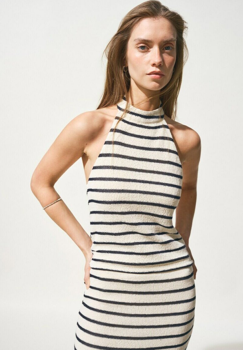 Femme aux longs cheveux châtain clair portant une robe blanche sans manches à rayures horizontales noires, debout les mains sur les hanches devant un fond uni.