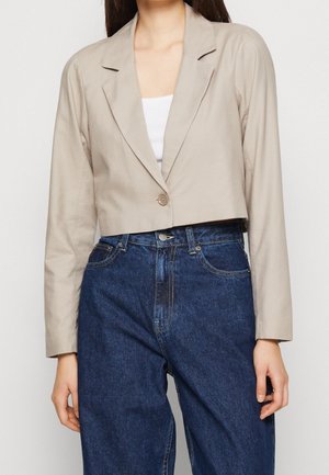 Kvinde iført en kort beige blazer med en enkelt knap over en hvid top, kombineret med højttaljede mørkeblå jeans.