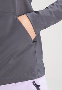 Mano de una persona insertada en el bolsillo delantero de una chaqueta gris de manga larga, mostrando la textura de la tela y los detalles de la costura.