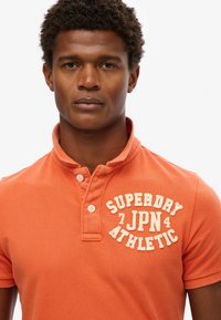 Pomarańczowa koszulka polo o teksturowanej tkaninie, z kołnierzykiem zapinanym na dwa guziki oraz białym, haftowanym logo "SUPERDRY JPN ATHLETIC" na piersi.
