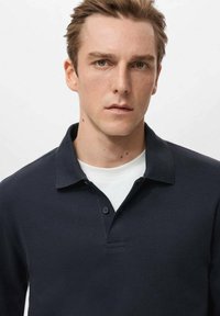 Temno modra polo majica z ovratnikom, dvo gumbnim zapenjanjem in kratkimi rokavi, nošena preko bele majice s krožnim izrezom. Gladek tekstura tkanine.