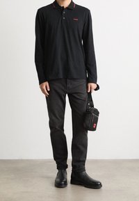 Schwarzes Langarmpolo mit roten Kragenakzenten, getragen zu dunklen Jeans und schwarzen Stiefeletten. Eine kleine schwarze Tasche wird in der rechten Hand gehalten.