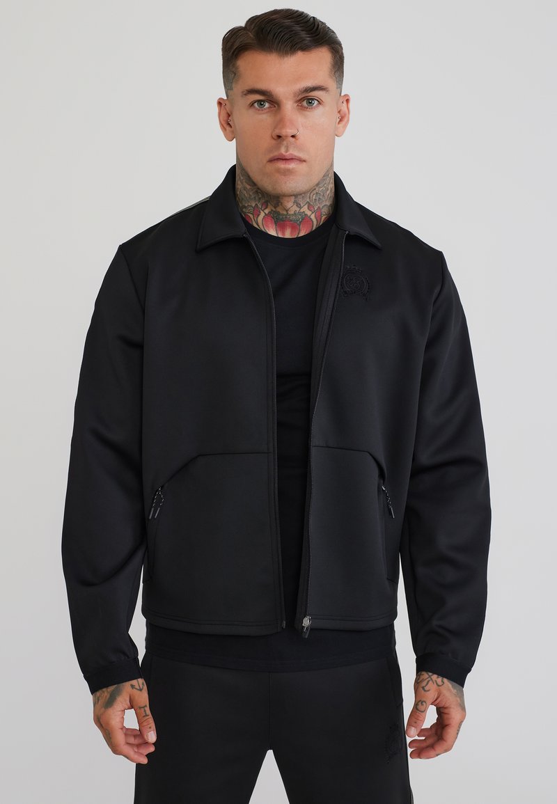 Man with neck and hand tattoos wearing a black zip-up jacket and matching black pants against a plain light background.