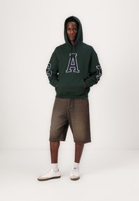 Mørkegrønn hettegenser med store hvite bokstaven "A", lange ermer og frontlomme. Kombinert med brune denimshorts og hvite joggesko.